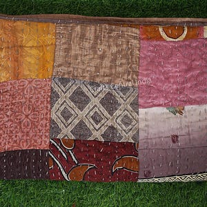 Könnte beinhalten: Ein Patchwork-Textil mit verschiedenen Stoffen in Braun-, Orange- und Rosatönen. Das rechteckige Stück besteht aus mehreren Quadraten, die mit weißem Faden zusammengenäht sind. Das Textil hat ein rustikales, handwerkliches Aussehen.