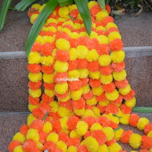 May include: A garland of yellow and orange fabric marigold flowers. The garland is made of individual pom-poms and is strung together with green string. The garland is lying on a set of brown stone steps.