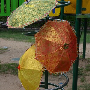 May include: Three colorful umbrellas with gold accents and fringe trim are displayed on a metal stand. The umbrellas are green, red, and yellow.