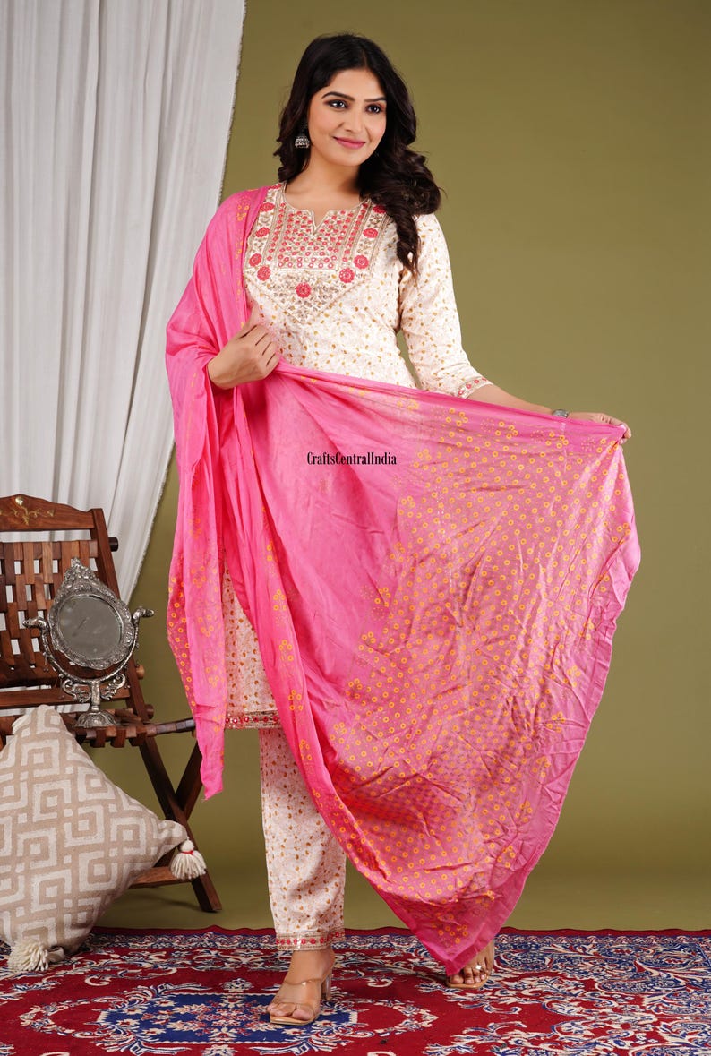 May include: A white cotton salwar kameez with pink and gold embroidery. The outfit includes a long-sleeved top, trousers, and a pink dupatta with a floral print.