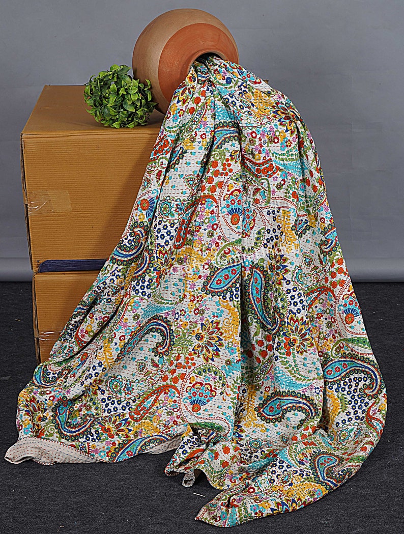 May include: A white kantha quilt with a paisley pattern in shades of blue, green, orange, and red. The quilt is draped over a cardboard box and a terracotta pot.