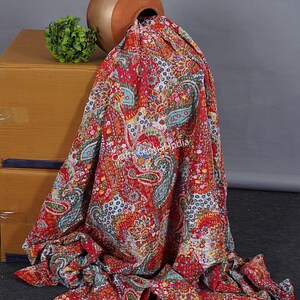 May include: A red, white and blue kantha quilt with a paisley pattern. The quilt is draped over a brown clay pot and a small green plant.