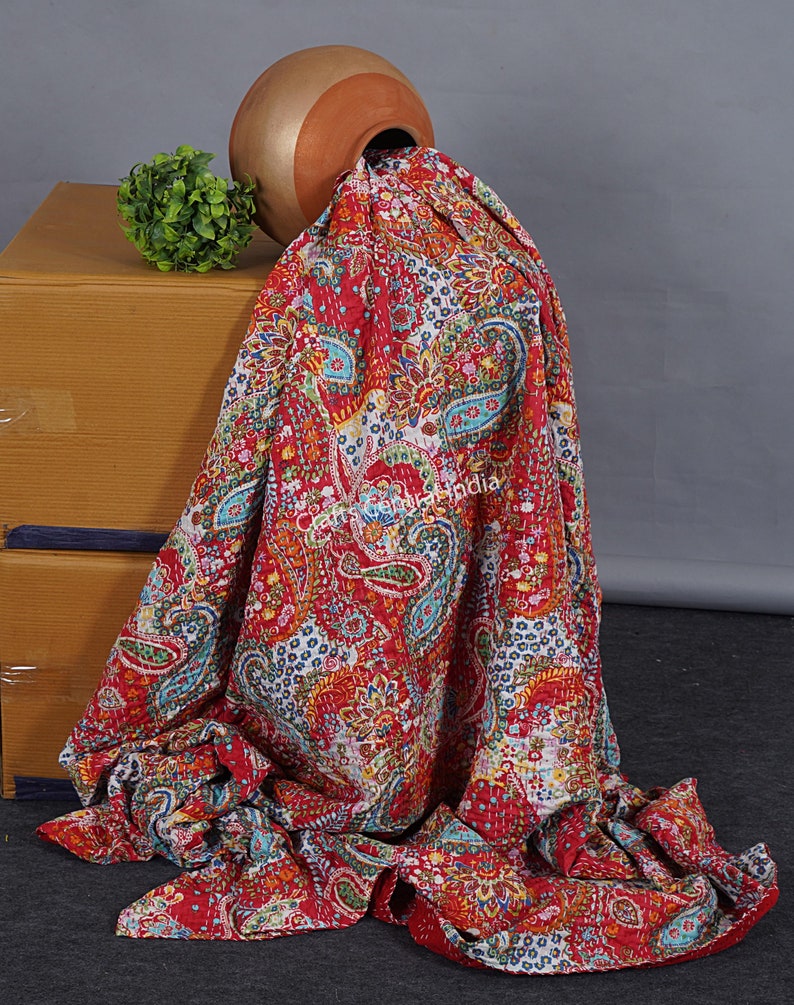 May include: A red, white and blue kantha quilt with a paisley pattern. The quilt is draped over a brown clay pot and a small green plant.