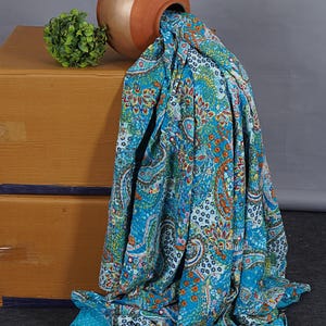 Könnte beinhalten: Eine leuchtend blaue Baumwolldecke mit einem mehrfarbigen Paisley- und Blumenmuster sowie feinen Steppnähten. Der Stoff fließt aus einem runden, terrakottafarbenen Tontopf und zeigt eine lebendige Mischung aus orangefarbenen, grünen und weißen Details.