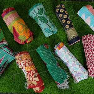 Puede incluir: Una colecci&oacute;n de diez colchas kantha coloridas enrolladas y dispuestas sobre una superficie de c&eacute;sped artificial verde. Las colchas est&aacute;n hechas de saris reciclados y presentan varios patrones y colores, incluyendo verde, azul, rojo, amarillo y blanco.