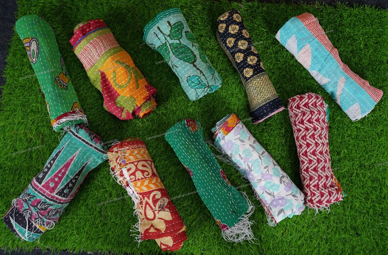 Puede incluir: Una colecci&oacute;n de diez colchas kantha coloridas enrolladas y dispuestas sobre una superficie de c&eacute;sped artificial verde. Las colchas est&aacute;n hechas de saris reciclados y presentan varios patrones y colores, incluyendo verde, azul, rojo, amarillo y blanco.