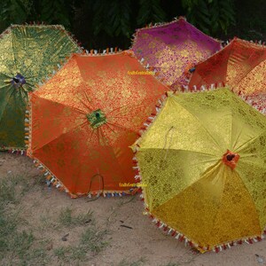 May include: Five colorful umbrellas with intricate patterns and gold accents. The umbrellas are made of fabric and have a fringed trim. They are arranged in a semi-circle on a dirt surface.