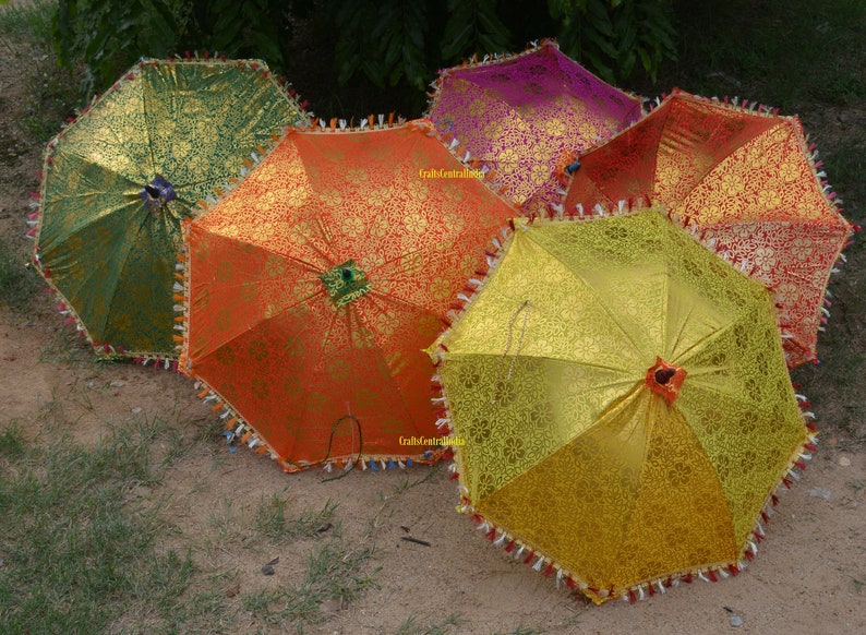 May include: Five colorful umbrellas with intricate patterns and gold accents. The umbrellas are made of fabric and have a fringed trim. They are arranged in a semi-circle on a dirt surface.