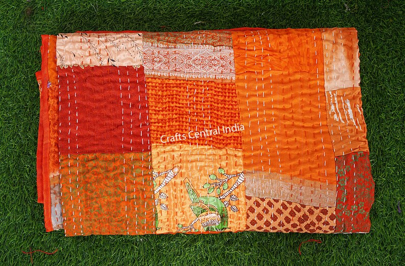 May include: A patchwork quilt made from colorful fabric scraps in shades of orange, red, and brown. The quilt is hand-stitched with white thread and has a unique, rustic look. The quilt is labeled "Crafts Central India".