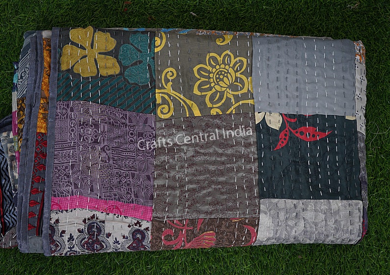 May include: A patchwork quilt with a variety of floral and geometric patterns in shades of gray, blue, pink, yellow, and red. The quilt is made of recycled saris and is hand-stitched with white thread. "Crafts Central India" is printed on the quilt.