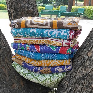 May include: A stack of eight colorful kantha quilts, made from recycled saris, with various floral and geometric patterns. The quilts are folded and stacked on a tree trunk.