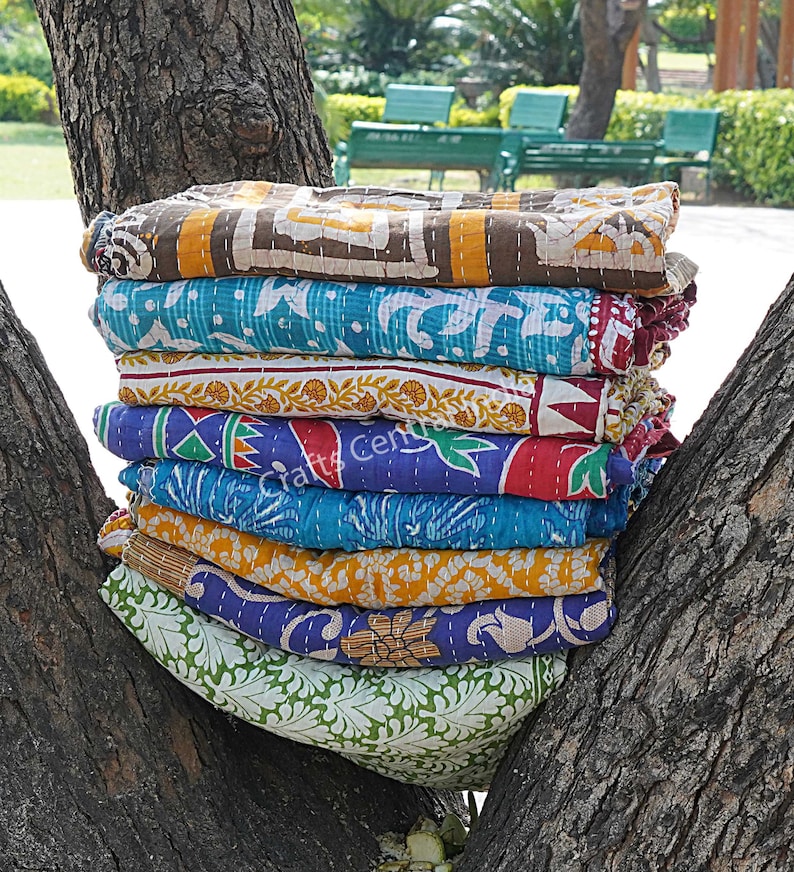 May include: A stack of eight colorful kantha quilts, made from recycled saris, with various floral and geometric patterns. The quilts are folded and stacked on a tree trunk.