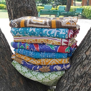 Könnte beinhalten: Ein Stapel von acht bunten Kantha-Steppdecken, hergestellt aus recycelten Saris, mit verschiedenen floralen und geometrischen Mustern. Die Decken sind gefaltet und auf einem Baumstamm gestapelt.
