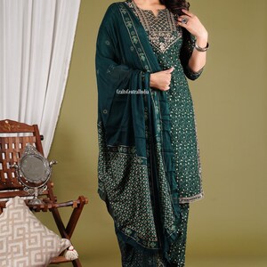 May include: A green and gold patterned salwar kameez with a matching dupatta. The outfit features a floral print and gold embroidery. The model is wearing the outfit and standing on a red and blue patterned rug.