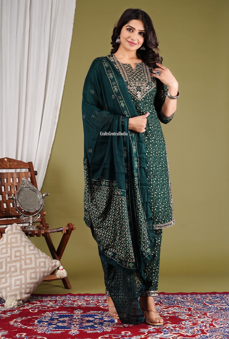 May include: A green and gold patterned salwar kameez with a matching dupatta. The outfit features a floral print and gold embroidery. The model is wearing the outfit and standing on a red and blue patterned rug.