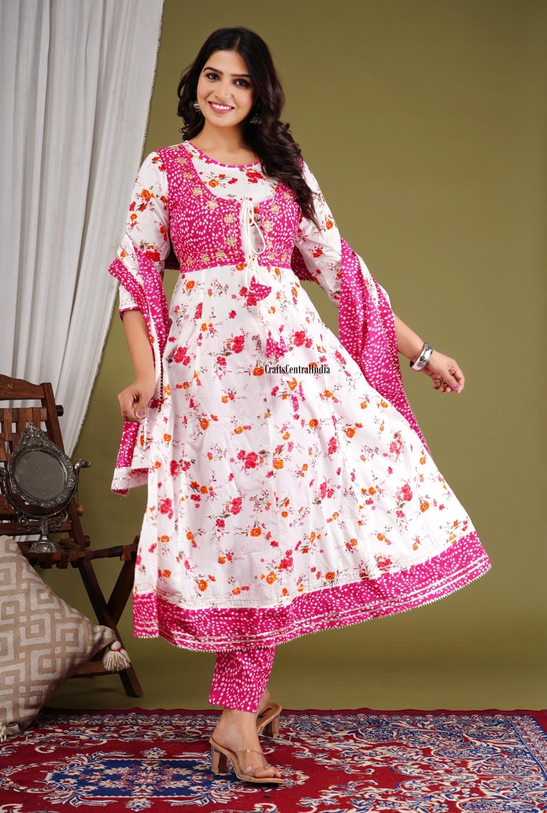 May include: A white and pink floral print kurta with a pink embroidered bodice and matching pants. The kurta has a tie-up neckline and a pink dupatta with white polka dots.