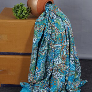May include: A turquoise blue kantha quilt with a paisley pattern and floral accents. The quilt is draped over a terracotta pot, which is resting on a wooden box.