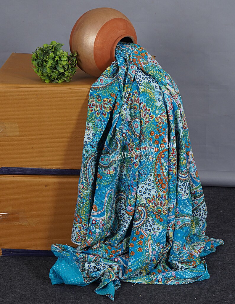 May include: A turquoise blue kantha quilt with a paisley pattern and floral accents. The quilt is draped over a terracotta pot, which is resting on a wooden box.
