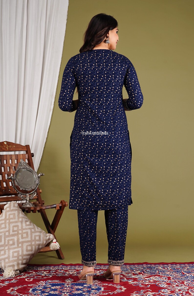 May include: A navy blue printed kurta and palazzo set with gold accents. The set features a floral print and a round neckline. The model is wearing the set and standing on a red patterned rug.