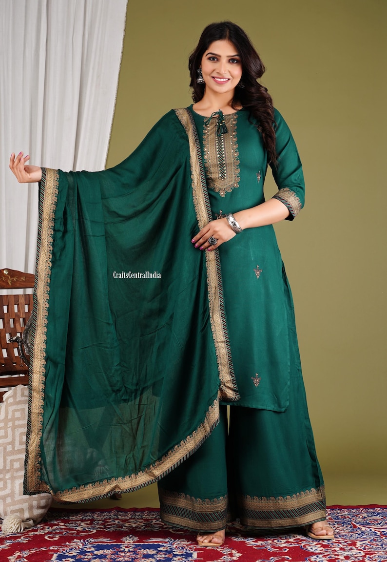 May include: A teal green kurta with gold embroidery and a matching dupatta. The kurta has a round neckline and three-quarter sleeves. The dupatta has a gold border and is draped over the shoulder. The outfit is perfect for a special occasion.