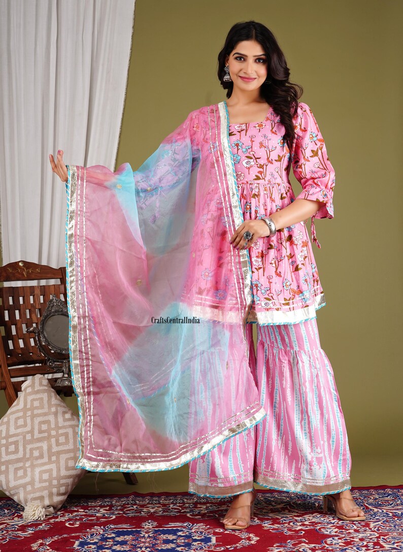 Op de afbeelding: Een roze en blauwe bedrukte jurk met een bijpassende dupatta. De jurk heeft een bloemenprint en een zwierige rok. De dupatta is doorschijnend en heeft een zilveren rand. De jurk wordt gedragen door een vrouw met lang donker haar. De tekst "CraftsCentralIndia" is zichtbaar in de afbeelding.