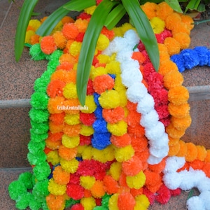 Könnte beinhalten: Eine bunte Girlande aus künstlichen Blumen mit leuchtend gelben, orangefarbenen, roten, blauen, grünen und weißen Pompons. Die Girlande ist aus Stoff und eignet sich perfekt zum Dekorieren für eine Party oder einen besonderen Anlass.