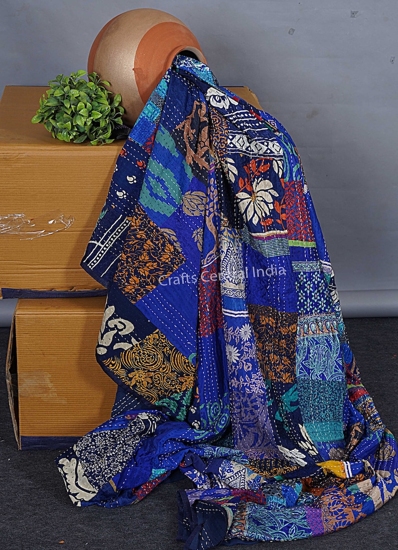 May include: A blue kantha quilt with a patchwork design featuring various floral and geometric patterns. The quilt is made of recycled saris and is hand-stitched. The quilt is draped over a box and a clay pot with a green plant.