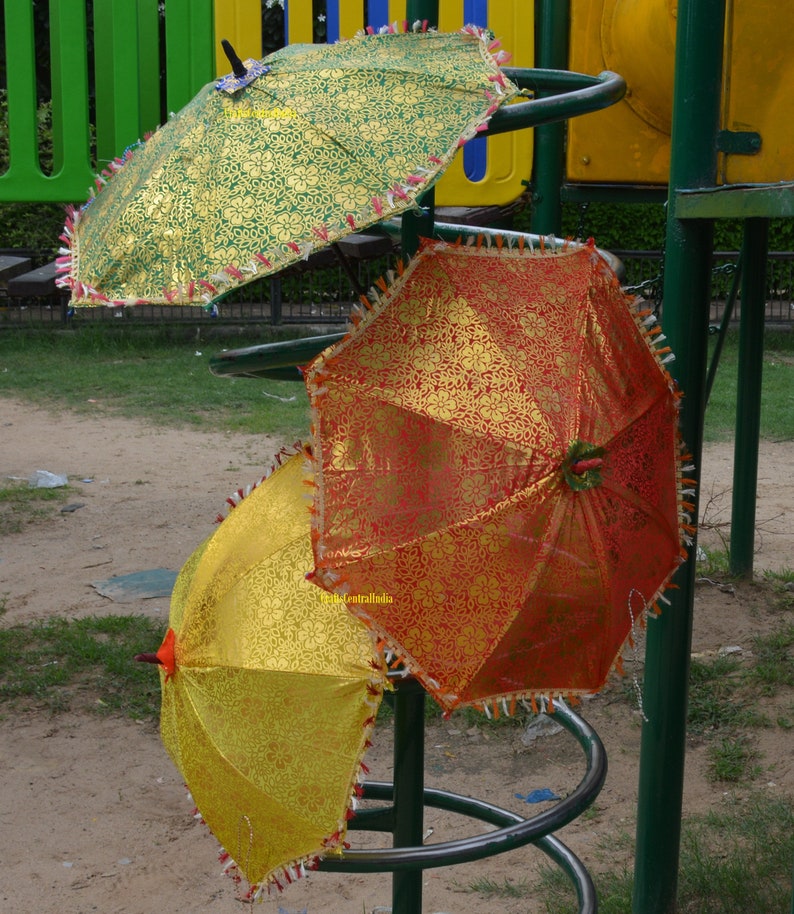 May include: Three colorful umbrellas with gold accents and fringe trim are displayed on a metal stand. The umbrellas are green, red, and yellow.