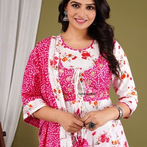 May include: A white and pink floral print dress with a matching pink dupatta. The dress has a white embroidered neckline and a tie-up detail at the front. The dupatta has a silver border and a white tassel.