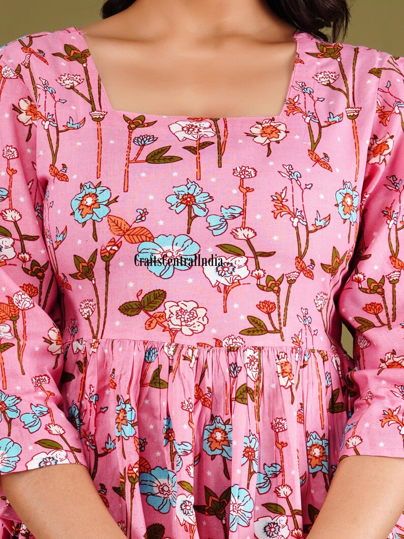 Op de afbeelding: Een roze katoenen jurk met een vierkante halslijn en een bloemenprint. De jurk heeft een geplooide rok en driekwart mouwen.