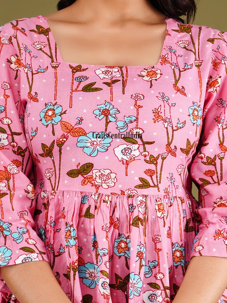 Op de afbeelding: Een roze katoenen jurk met een vierkante halslijn en een bloemenprint. De jurk heeft een geplooide rok en 3/4 mouwen.
