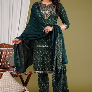 May include: A woman is wearing a green printed salwar kameez with gold embroidery. The outfit includes a matching dupatta. The outfit is a traditional Indian garment.