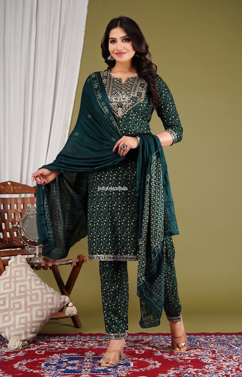 May include: A woman is wearing a green printed salwar kameez with gold embroidery. The outfit includes a matching dupatta. The outfit is a traditional Indian garment.