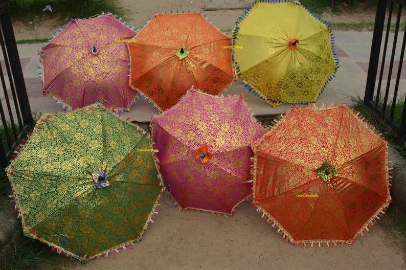 May include: Six colorful umbrellas with gold accents and fringe trim. The umbrellas are arranged in a semi-circle on a brown surface.