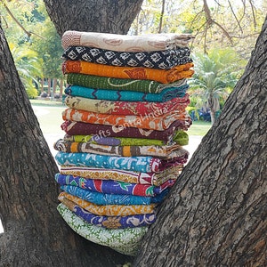 May include: A stack of colorful kantha quilts, made from recycled saris, are displayed between two tree trunks. The quilts are layered in a variety of patterns and colors, including orange, green, blue, red, and brown. The text "Crafts Central India" is visible on one of the quilts.