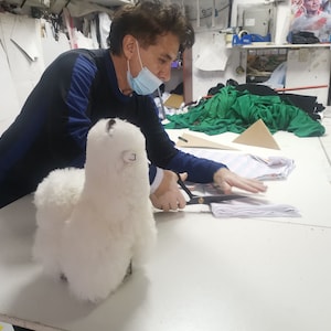K&ouml;nnte beinhalten: Eine Person mit einer Gesichtsmaske schneidet Stoff mit einer Schere, w&auml;hrend ein wei&szlig;es, flauschiges Alpaka-Spielzeug auf dem Tisch liegt.