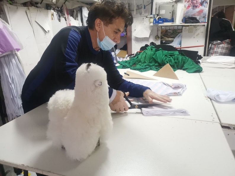 K&ouml;nnte beinhalten: Eine Person mit einer Gesichtsmaske schneidet Stoff mit einer Schere, w&auml;hrend ein wei&szlig;es, flauschiges Alpaka-Spielzeug auf dem Tisch liegt.