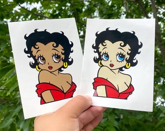 Puede incluir: Dos pegatinas rectangulares con un personaje de dibujos animados de pelo negro, pendientes de aro dorados y un vestido rojo con hombros descubiertos. Una pegatina muestra al personaje con piel bronceada y ojos marrones, y la otra con piel clara y ojos azules.