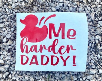 Puede incluir: Una pegatina rectangular blanca con texto rojo y un gráfico de pato rojo. El texto dice "Me harder DADDY!" en una fuente cursiva juguetona. La pegatina está colocada sobre un lecho de pequeñas piedras grises.