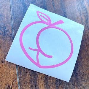 May include: Pink peach sticker with a simple outline design. The sticker is on a white background, with a wooden surface in the background.