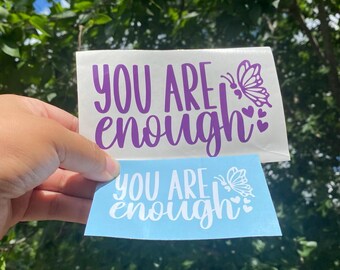 Puede incluir: Dos pegatinas rectangulares de vinilo con el texto "YOU ARE enough" en una fuente manuscrita estilizada. La pegatina superior tiene texto morado sobre fondo blanco, mientras que la inferior tiene texto blanco sobre fondo azul claro, ambas con mariposa.
