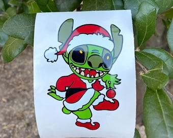 May include: A green cartoon character wearing a Santa hat and red and white Santa suit. The character is holding a red ornament. The sticker is on a white background with green leaves in the background.