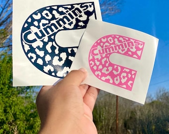 Puede incluir: Dos calcomanías de vinilo blanco con la palabra "Cummins". Una es azul marino con estampado de leopardo, la otra rosa con estampado de leopardo. Las calcomanías se sostienen contra un cielo azul. Para un vehículo.