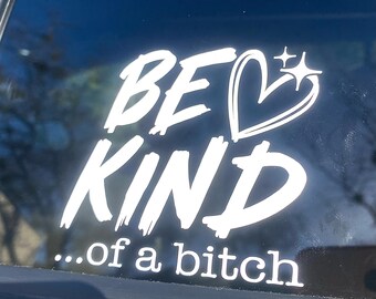 Puede incluir: Calcomanía de vinilo blanco con las palabras "BE KIND" encima de "...of a bitch" en una fuente en negrita y con trazos de pincel. Un corazón y un gráfico de estrella están junto a la palabra "BE". La calcomanía está aplicada a una superficie oscura.