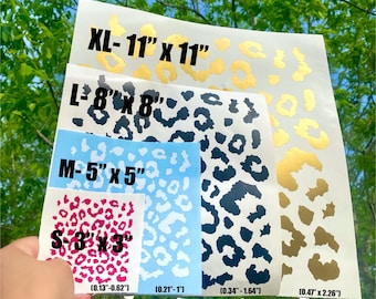 Puede incluir: Hojas de calcomanías con estampado de leopardo en varios tamaños, incluyendo XL (28 cm x 28 cm), L (20 cm x 20 cm), M (13 cm x 13 cm) y S (8 cm x 8 cm). Las calcomanías presentan manchas de leopardo doradas, negras y rosas. La imagen también muestra el tamaño de las manchas.