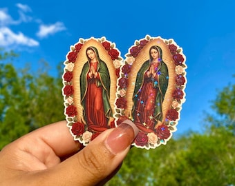 Puede incluir: Dos pegatinas que representan una figura religiosa con un vestido rojo y una capa verde, rodeada de rosas rojas y blancas. Las pegatinas se sostienen contra un cielo azul brillante y follaje verde.