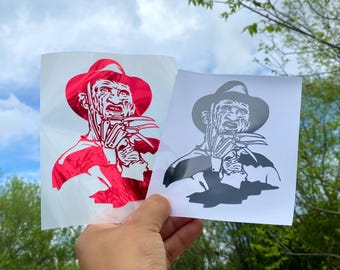 Puede incluir: Dos pegatinas con un personaje de película de terror. Una pegatina es roja y la otra en blanco y negro. El personaje lleva sombrero y camisa a rayas, con un guante con garras. Las pegatinas se sostienen contra un cielo azul.