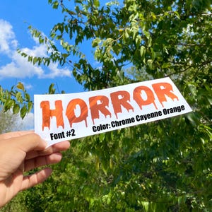 May include: A white sticker with the word "HORROR" in chrome cayenne orange, dripping font. The text "Font #2" and "Color: Chrome Cayenne Orange" are also on the sticker. The background is a blue sky and green foliage.
