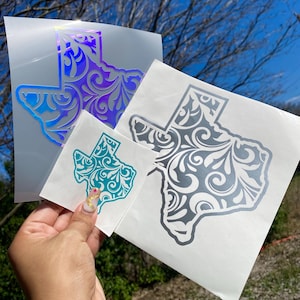 May include: Several Texas-shaped vinyl stickers in various sizes and colors. The stickers feature an ornate, swirling design within the state outline. Colors include iridescent, teal, and silver, with a clear background.