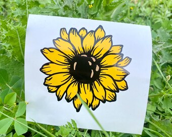 Puede incluir: Adhesivo de girasol amarillo y negro sobre fondo blanco. El girasol tiene un centro negro y pétalos amarillos. La pegatina está colocada sobre un lecho de hierba y hojas verdes.